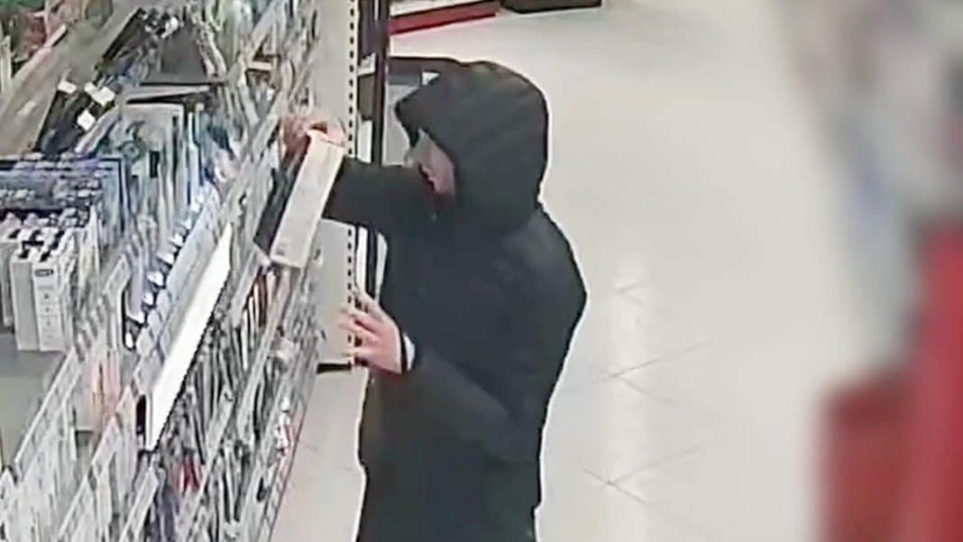 Mężczyzna w czarnej kurtce i kapturze sięga po produkt na p&oacute;łce w sklepie.