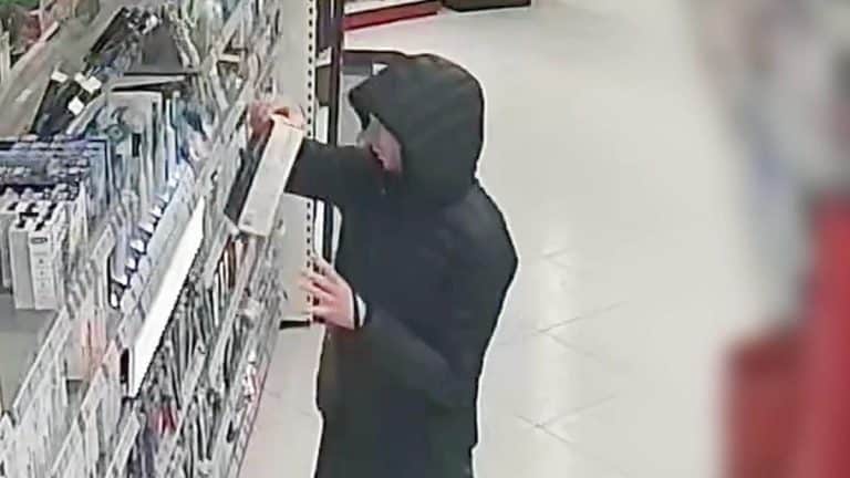 Mężczyzna w czarnej kurtce i kapturze sięga po produkt na p&oacute;łce w sklepie.