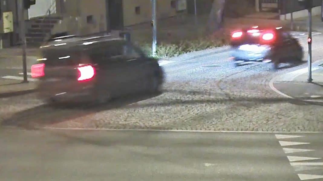 Zdjęcie z policyjnej interwencji, dwa samochody na ulicy w nocy, jeden z nich odjeżdża, w tle oświetlone budynki.