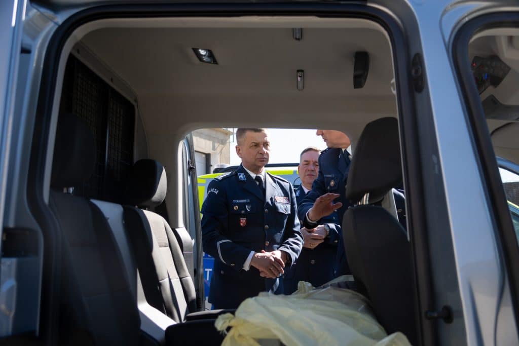 Policjanci prezentują nowe radiowozy na Mazurach, widok z wnętrza pojazdu, z ludźmi w mundurach na zewnątrz.