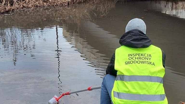 Pracownik inspekcji środowiska monitoruje zanieczyszczoną rzekę, na powierzchni widoczne plamy oleju.