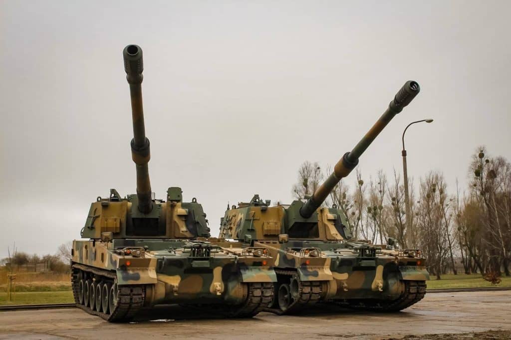 Dwa nowoczesne armatohaubice K9 Thunder na placu, z drzewami w tle, w pochmurny dzień.