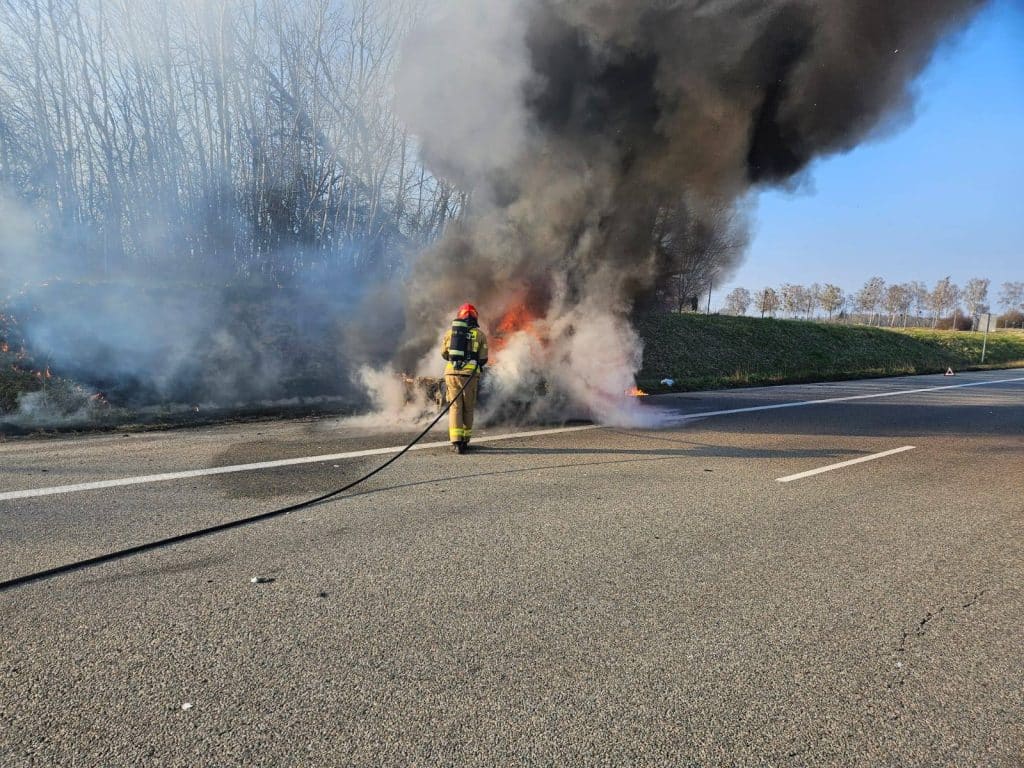 Strażak gasi pożar samochodu na drodze ekspresowej, w tle widoczne drzewa i niebo.