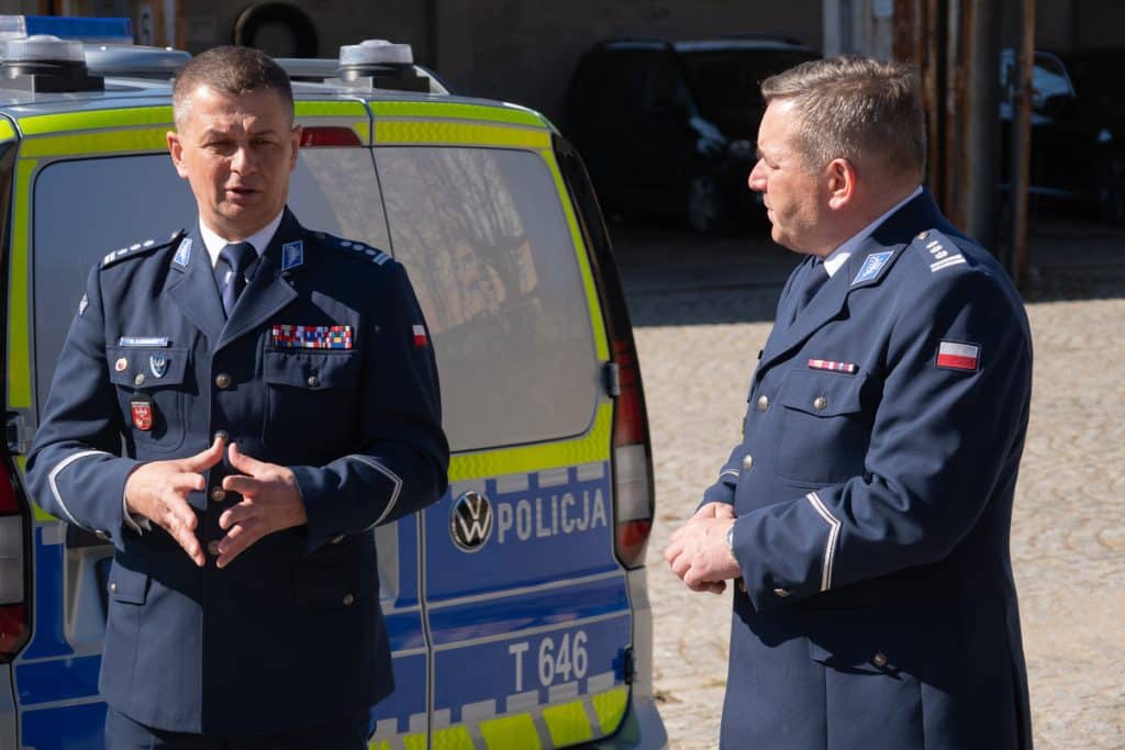 Dwa policyjni funkcjonariusze rozmawiają przy nowoczesnym radiowozie na Mazurach, w tle policyjny samoch&oacute;d.