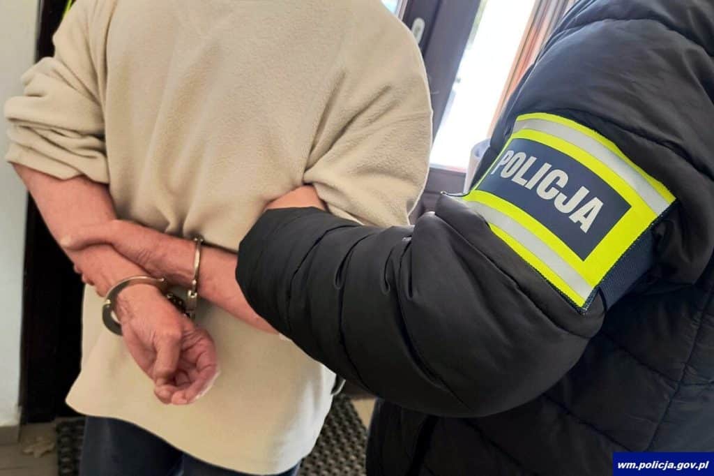 Policjant zatrzymuje osobę w rękawiczkach, w tle drzwi wejściowe, w pomieszczeniu.