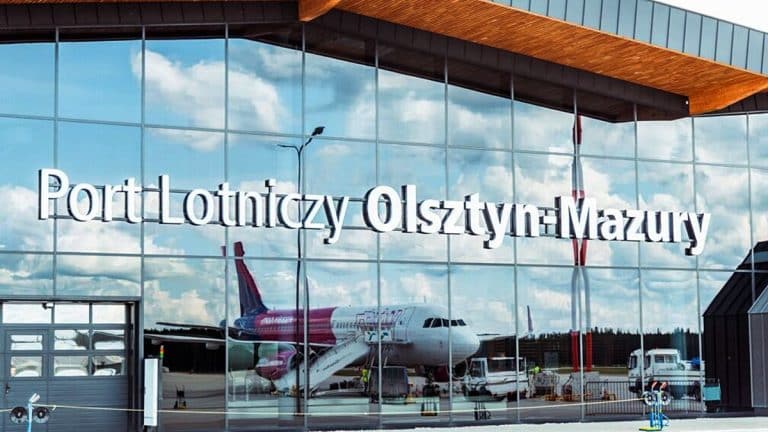 Widok na nowoczesny terminal lotniska Olsztyn-Mazury z samolotem na płycie lotniska, odbijający się w szklanych ścianach.