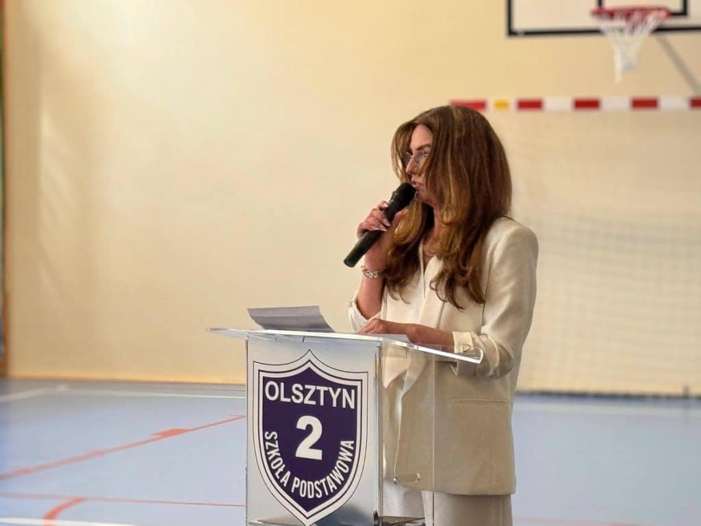 Kobieta przemawia do publiczności podczas otwarcia nowej hali sportowej w szkole podstawowej w Olsztynie.