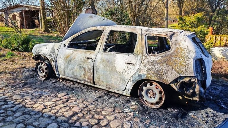 Spalony samoch&oacute;d osobowy na poboczu, uszkodzony i pozbawiony części, w tle drzewa i ogr&oacute;d.