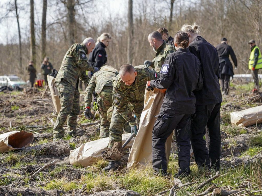 Grupa ludzi sadzi drzewa w lesie podczas wydarzenia na Mazurach, w tle drzewa i osoby w mundurach.