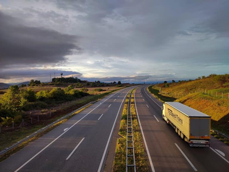 Pusty odcinek autostrady z ciężarówką i chmurami na niebie, otoczony zielonymi wzgórzami.
