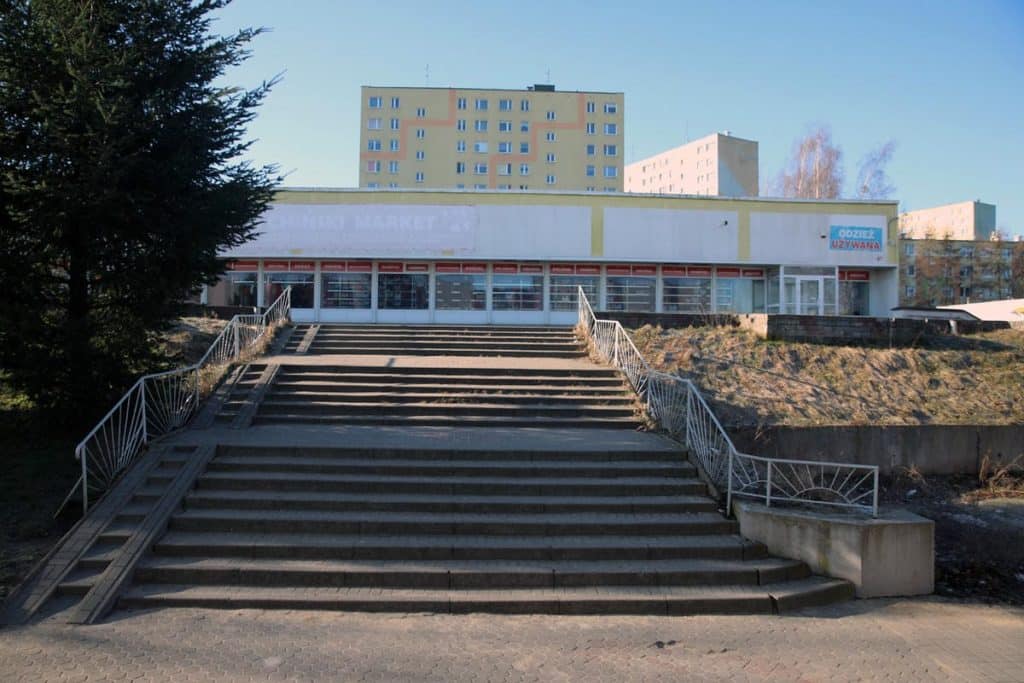 Budynek handlowy z wejściem i schodami, w tle bloki mieszkalne, na zdjęciu słoneczny dzień.