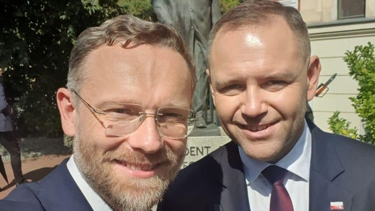 Dw&oacute;ch mężczyzn w garniturach na zewnątrz, uśmiechają się do kamery, w tle rośliny i budynek.