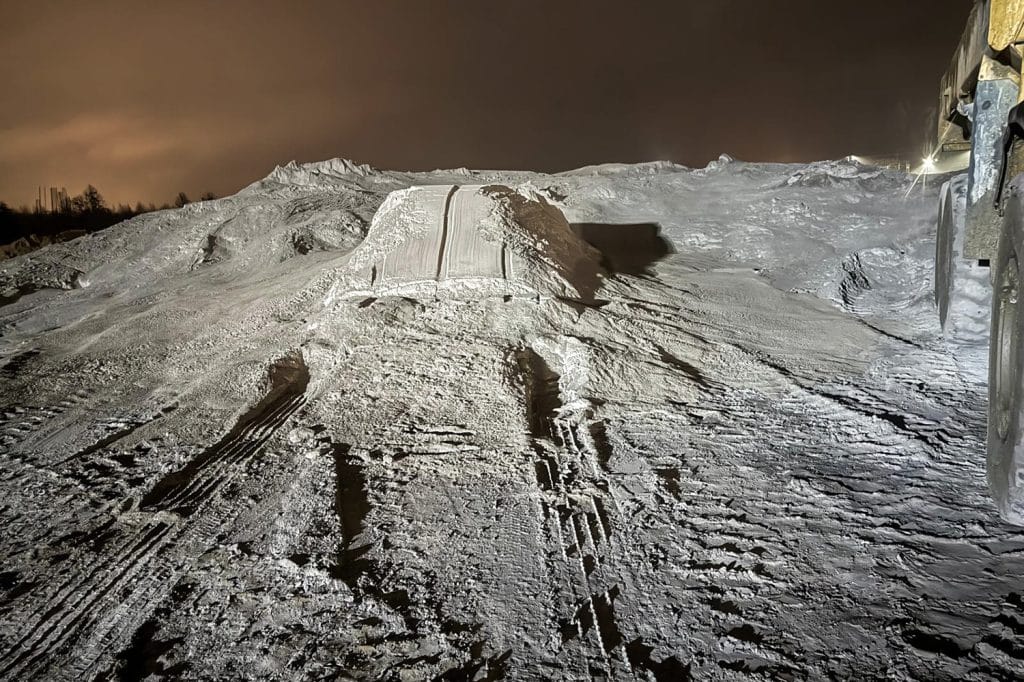Zdjęcie zaśnieżonego terenu nocą, ślady pojazd&oacute;w i śniegowe kopce, w tle ciemne niebo.