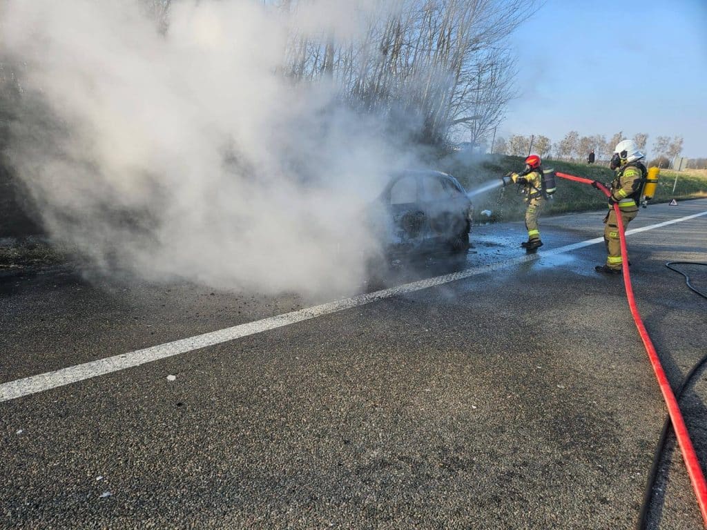 Strażacy gaszą pożar samochodu marki Audi na drodze ekspresowej S22, w tle widoczne drzewa i niebo.