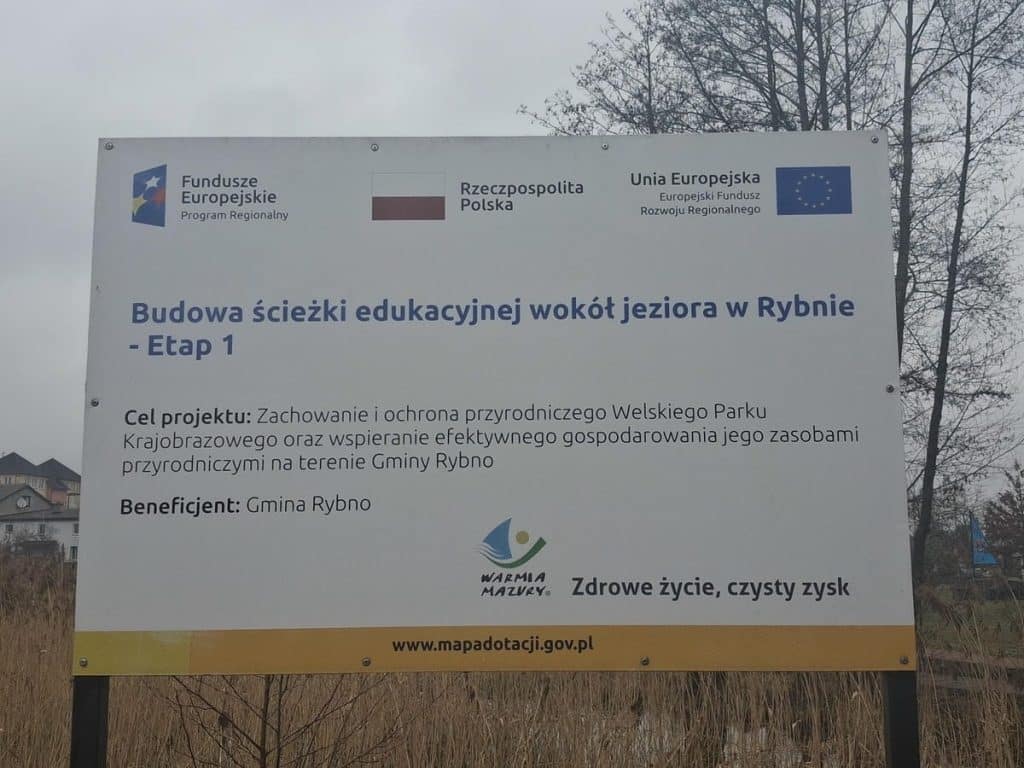 Tablica informacyjna o budowie ścieżki edukacyjnej wok&oacute;ł jeziora w Rybnie, z logami funduszy unijnych i gminy.
