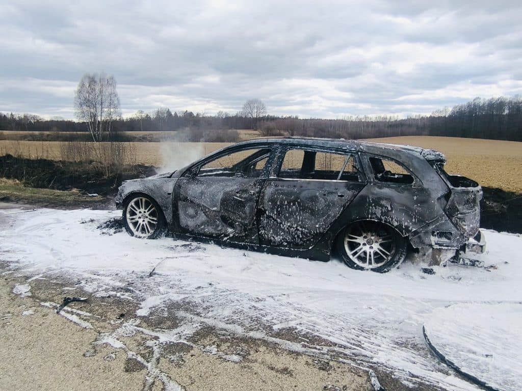 Spalony samoch&oacute;d na poboczu drogi, z dymem unoszącym się spod maski, na tle p&oacute;l i nieba.