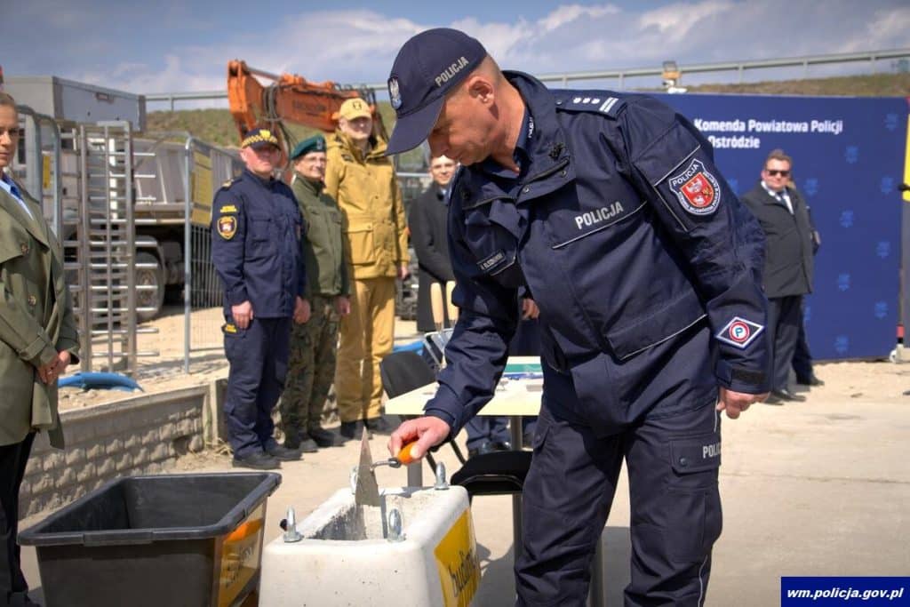 Policjant podczas ceremonii rozpoczęcia budowy nowej komendy w Ostr&oacute;dzie, w tle inni funkcjonariusze i symboliczny akt erekcyjny.