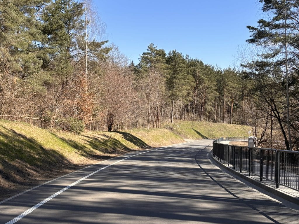 Nowa, szeroka droga w otoczeniu lasu, z barierkami po prawej stronie, na tle nieba i drzew.