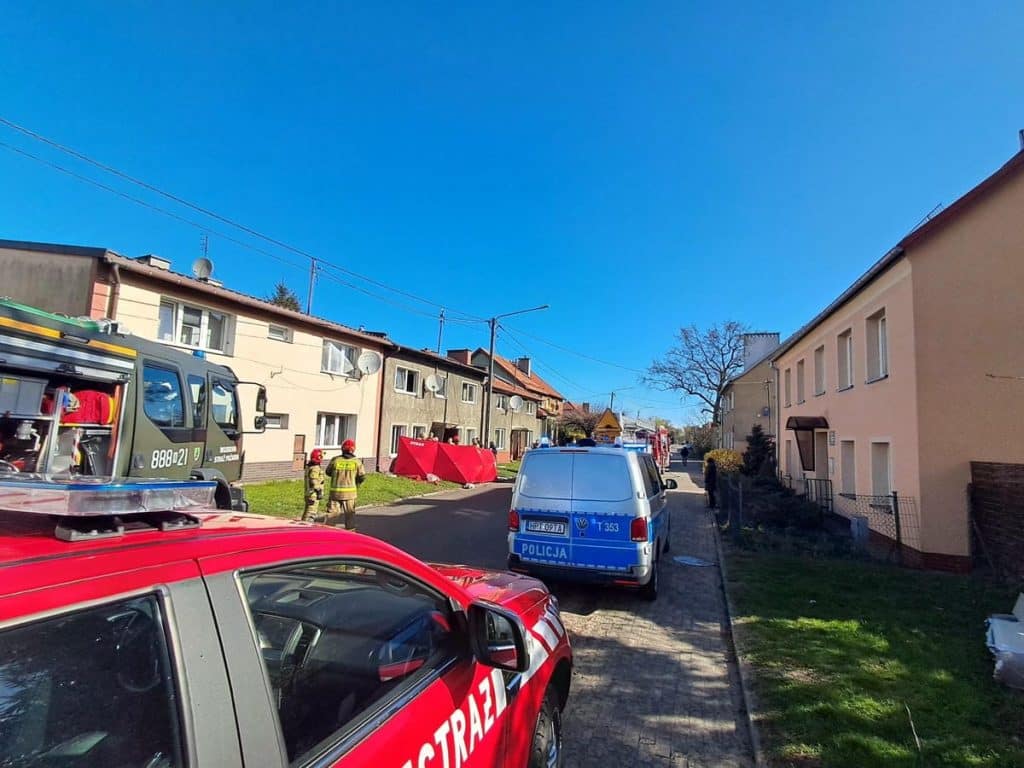 Strażacy w akcji przed blokiem mieszkalnym, wejście do mieszkania, pojazdy służb ratunkowych na ulicy.