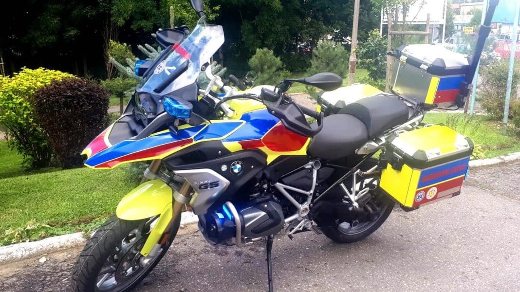 Motocykl ratunkowy z ż&oacute;łtymi i niebieskimi elementami, wyposażony w skrzynie na sprzęt, na tle zieleni i chodnika.