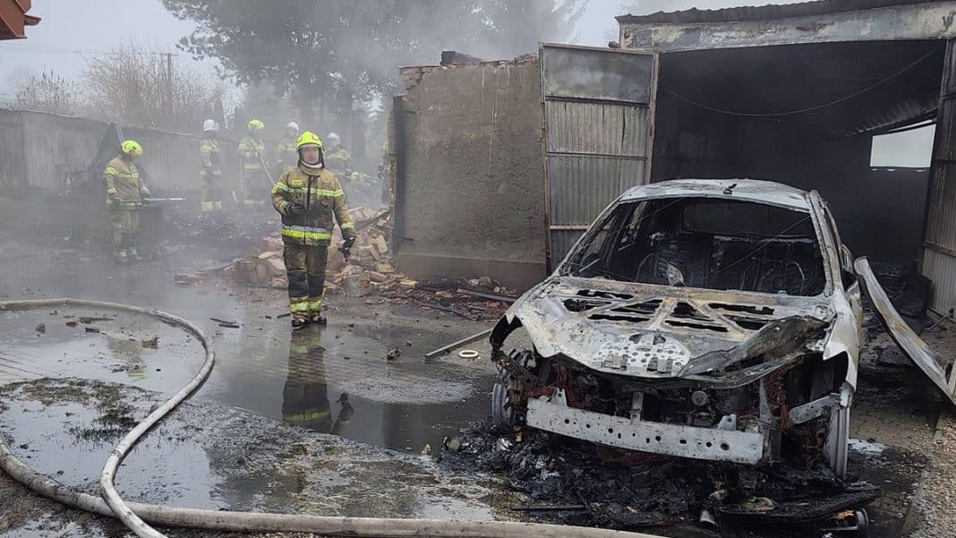 Zniszczony samoch&oacute;d i spalone wnętrze garażu po pożarze na Mazurach, strażacy w akcji w tle.