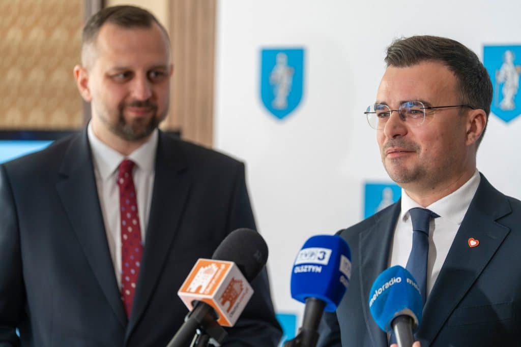 Dw&oacute;ch mężczyzn w garniturach podczas konferencji prasowej, za nimi herby miasta, mikrofony przed nimi.