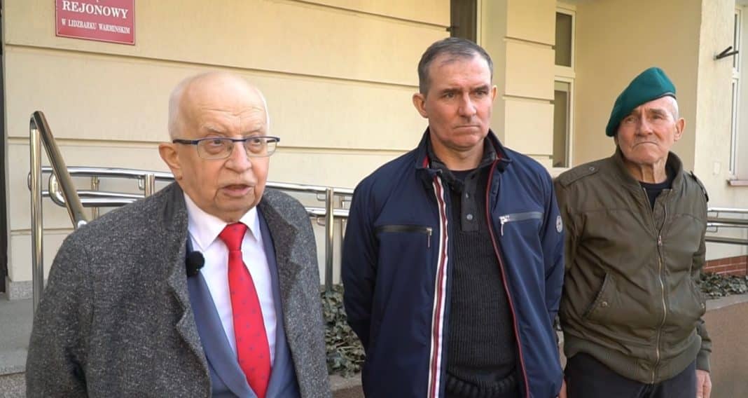 Trzech mężczyzn stojących na zewnątrz budynku, jeden z nich w garniturze i czerwonym krawacie.