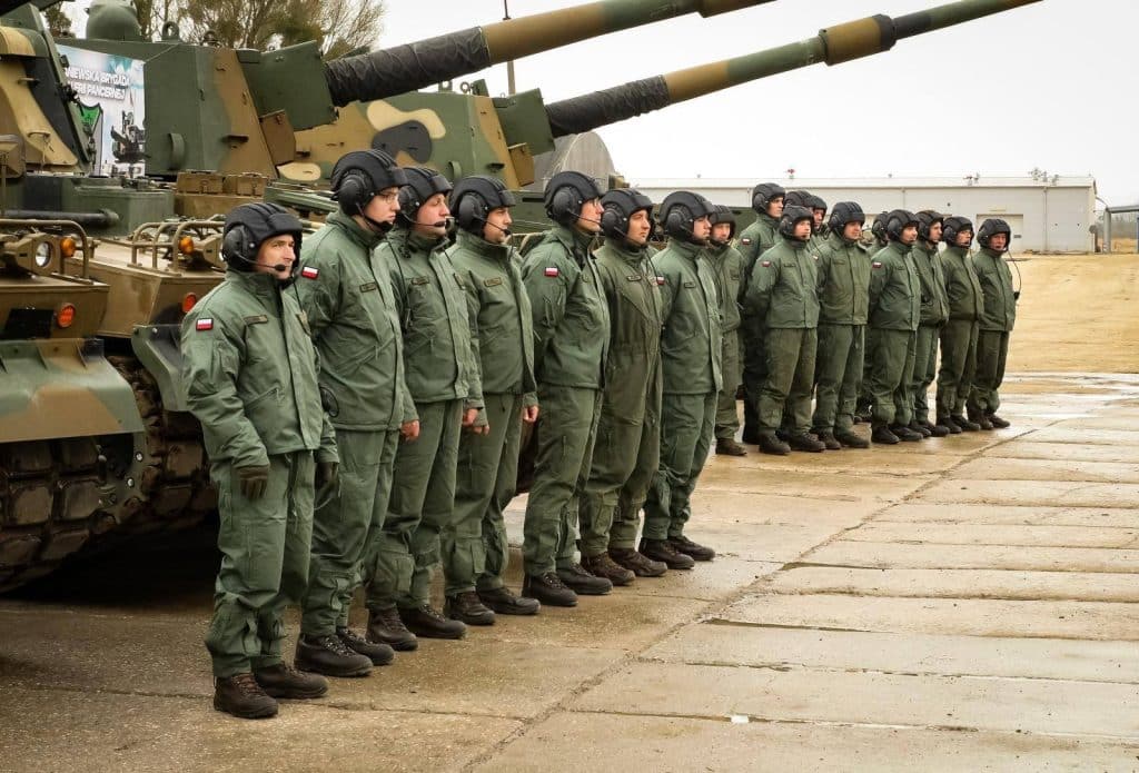 Żołnierze w hełmach i mundurach stojący obok armatohaubic na placu treningowym.