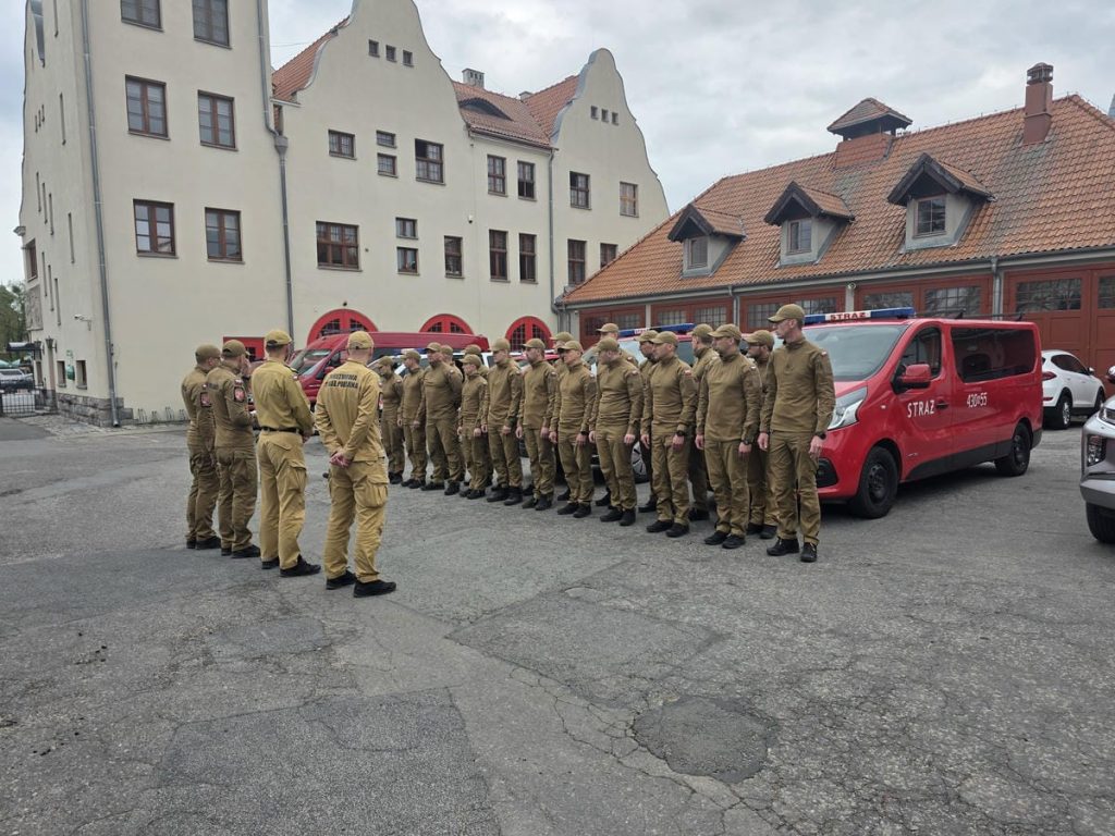 Grupa strażak&oacute;w w uniformach stojąca obok czerwonych pojazd&oacute;w na placu przed budynkiem.
