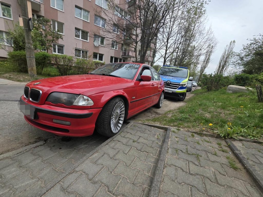 Czerwone BMW i policyjny samoch&oacute;d na ulicy w centrum Olsztyna, w tle blok mieszkalny i drzewa.