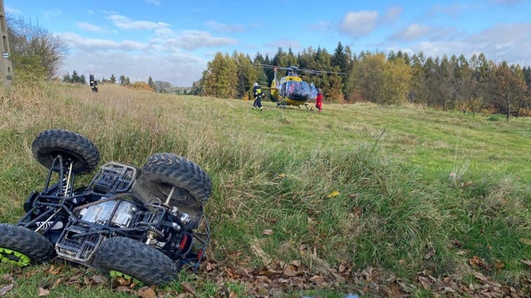 Rozbity quad na trawie, w tle śmigłowiec LPR i ratownicy na miejscu wypadku.