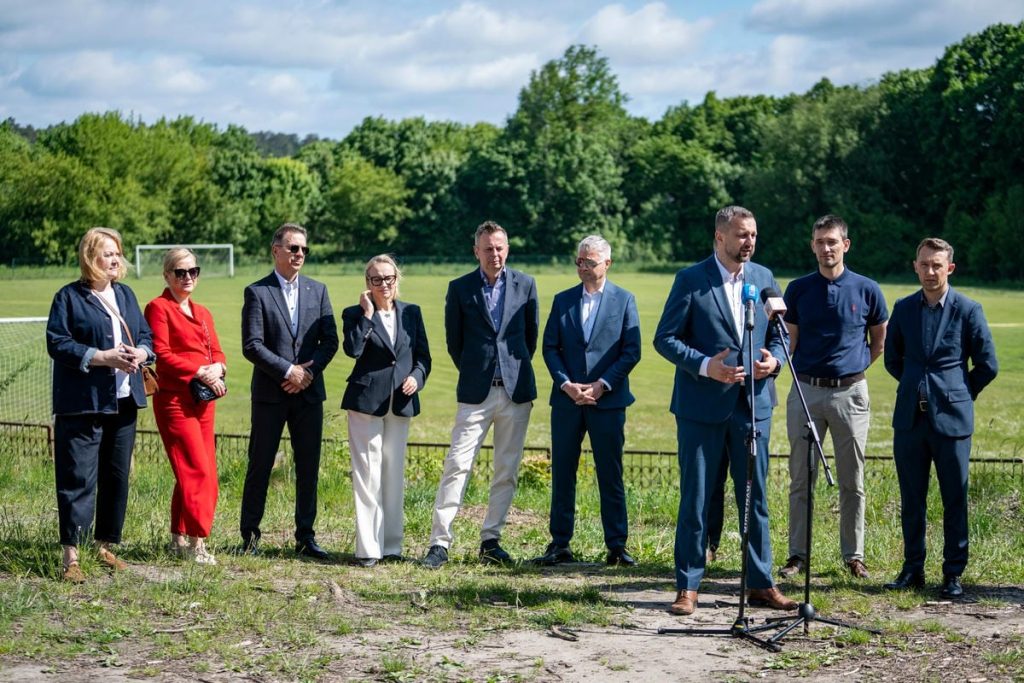 Grupa osób na otwarciu nowego obiektu sportowego na tle zielonego boiska i drzew.