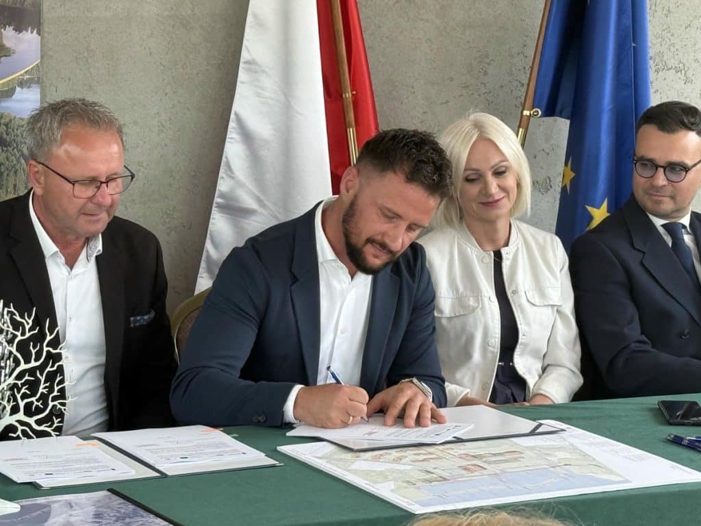 Osoby podpisujące umowę na budowę portu nad jeziorem Guzianka Wielka, z flagami w tle.