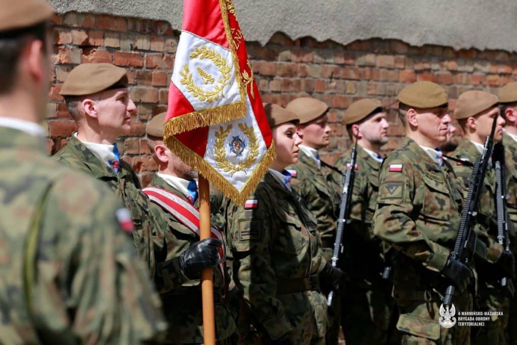 Żołnierze Obrony Terytorialnej w mundurach podczas uroczystości, trzymający flagę, na tle ceglanej ściany.