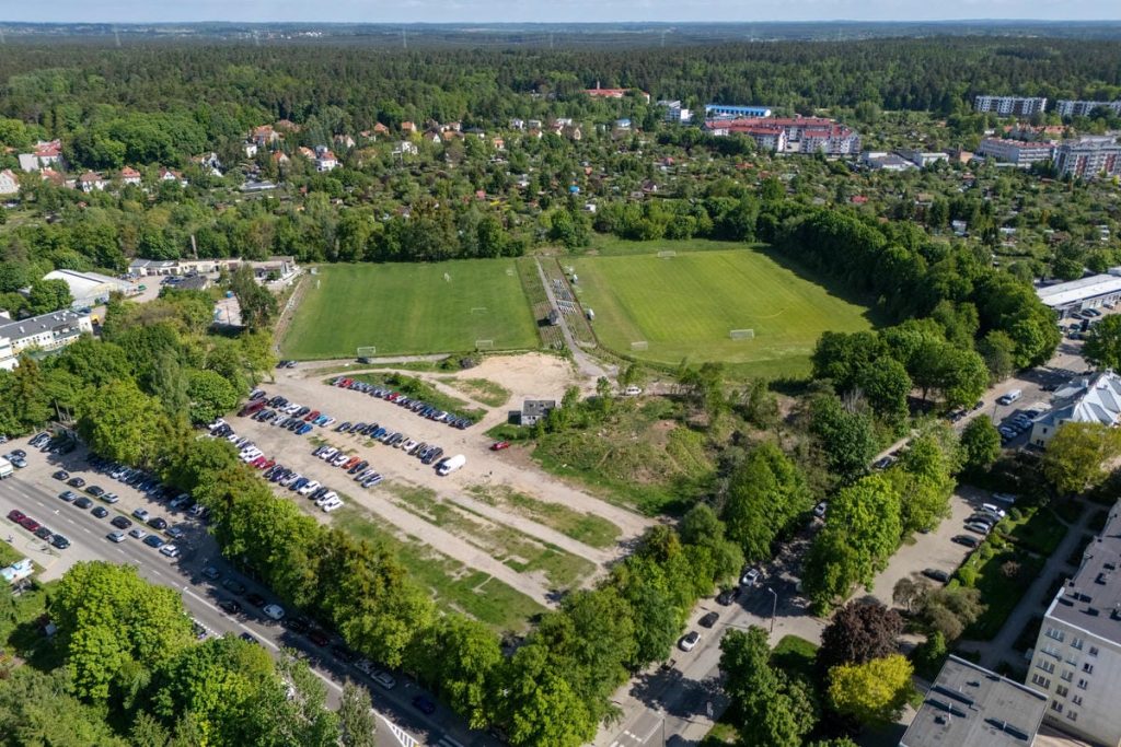 Widok na plac budowy nowoczesnego ośrodka piłkarskiego w Olsztynie, otoczony drzewami i istniejącymi boiskami.