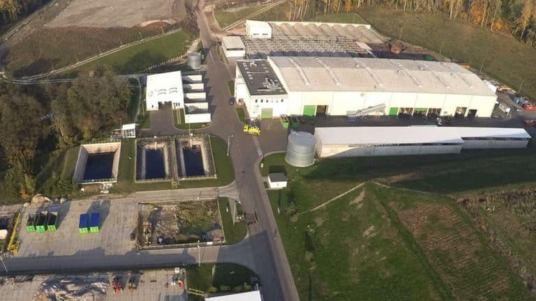 Widok na zakład utylizacji odpadów w Elblągu, w tym duże hale i zbiorniki na odpady, otoczony zielonym terenem.