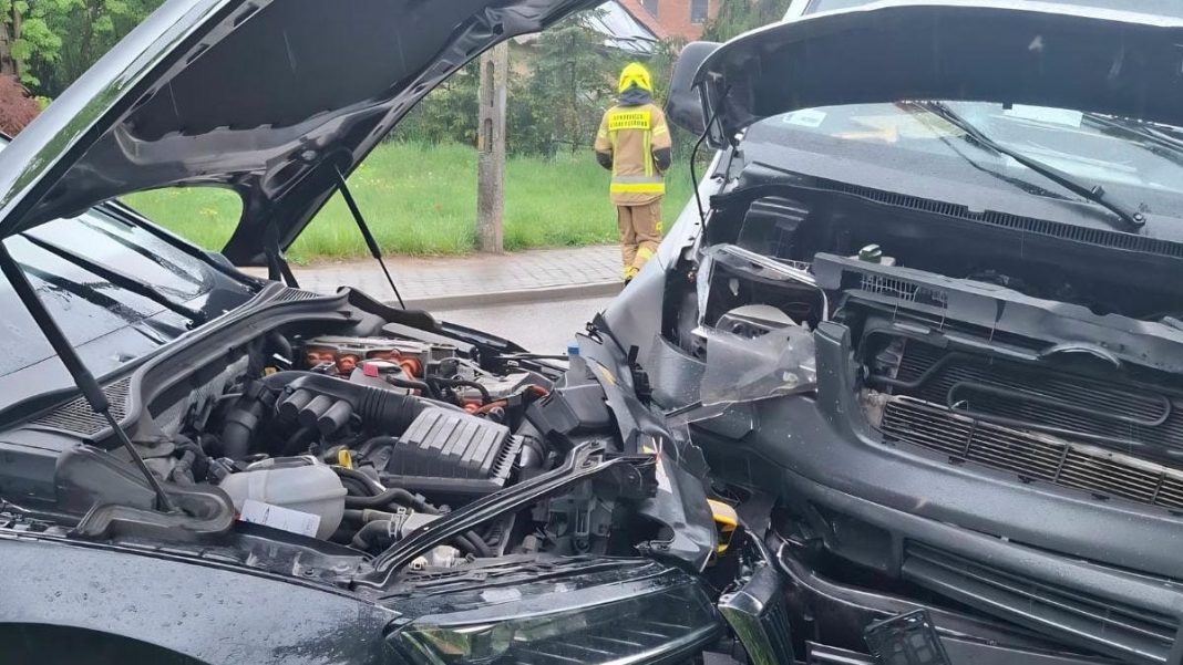 Zdjęcie przedstawia rozbity samoch&oacute;d hybrydowy i dostawczy po czołowym zderzeniu na drodze powiatowej, z widocznym strażakiem w tle.