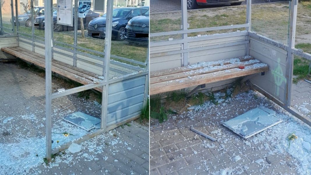 Zniszczony przystanek autobusowy z wybitymi szybami i rozbitymi elementami, na tle parkingu.