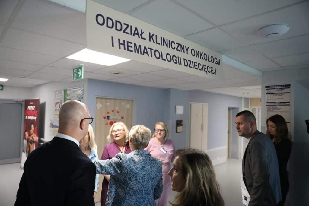 Grupa osób rozmawia w korytarzu szpitala, widoczny znak wskazujący na oddział onkologii dziecięcej.