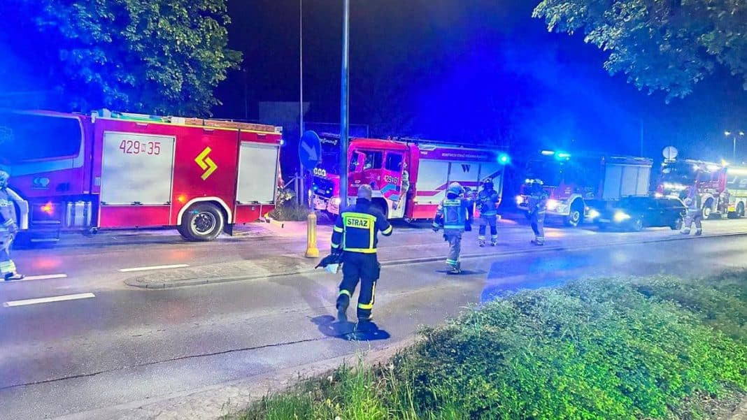 Straż pożarna interweniuje przy zakładzie przemysłowym, w tle pojazdy ratunkowe i rozświetlone światła.