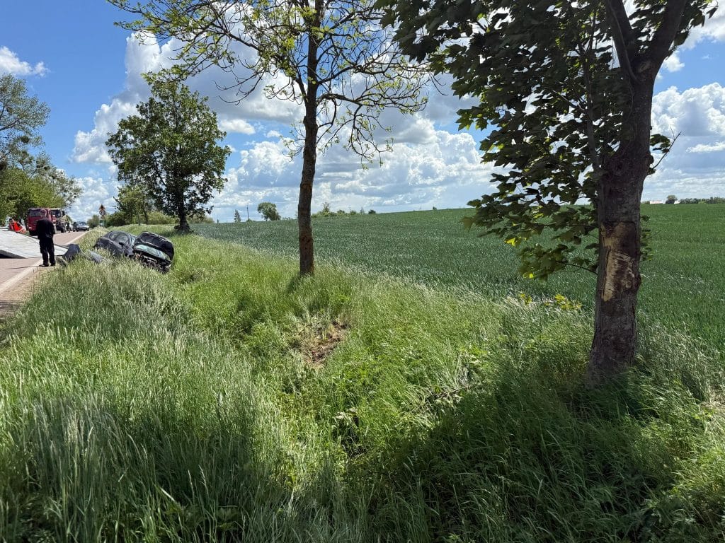Rozbity samochód marki Peugeot leży na poboczu drogi wojewódzkiej, w pobliżu drzew i zielonych pól, w rejonie Gałwunów.