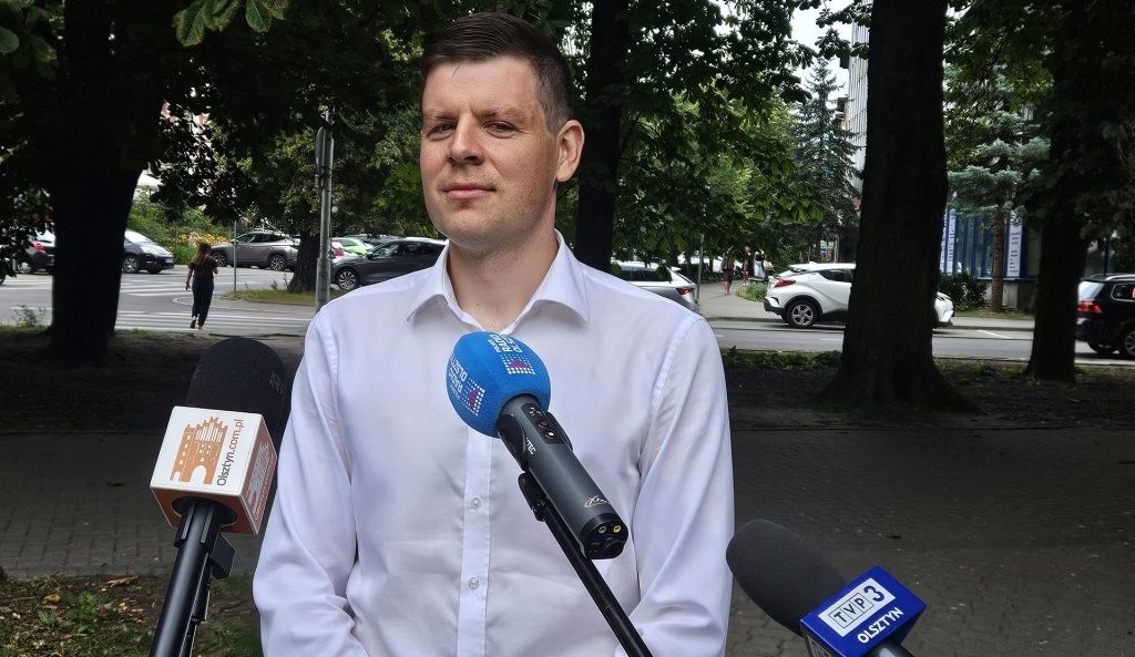 Mężczyzna w białej koszuli przemawia do medi&oacute;w na tle drzew i ulicy z samochodami.