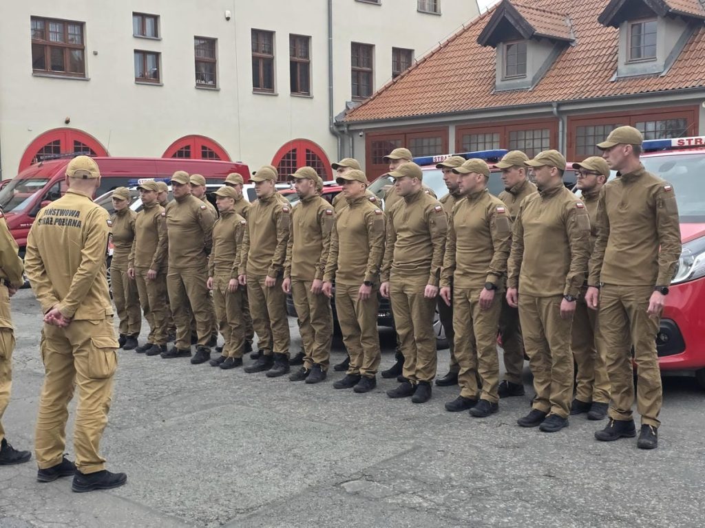 Grupa strażak&oacute;w w uniformach stojąca na placu, przygotowująca się do akcji gaśniczej.