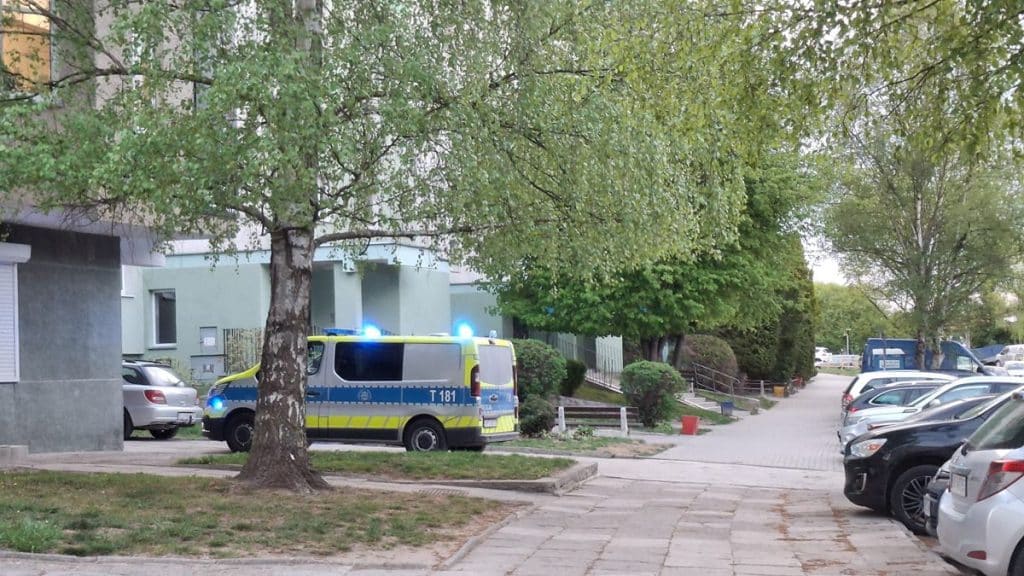 Widok na miejsce zdarzenia pod blokiem w Olsztynie, z radiowozem i zaparkowanymi samochodami.