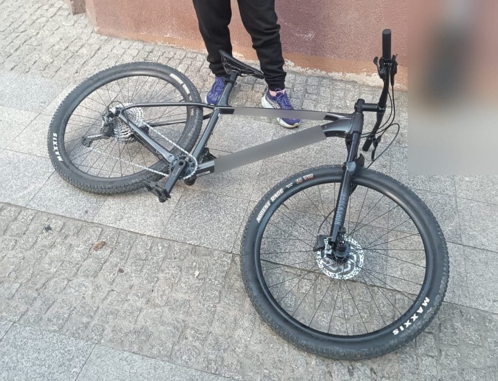 Rower leży na chodniku, obok stoi osoba w sportowym ubraniu, w tle ściana budynku.