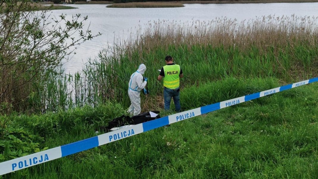 Policjanci i technik na brzegu jeziora, zabezpieczają miejsce makabrycznego odkrycia.