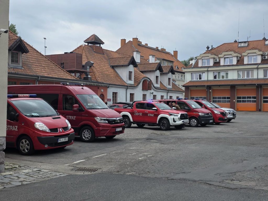 Grupa czerwonych samochod&oacute;w strażackich zaparkowanych na placu w mieście, w tle zabytkowe budynki z czerwonymi dachami.