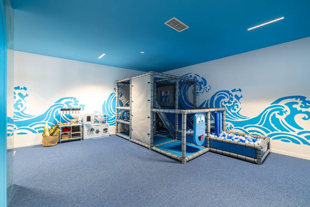 Wewnątrz plac zabaw dla dzieci z modułami do wspinaczki i basenem z piłkami, na ścianach mural z falami.