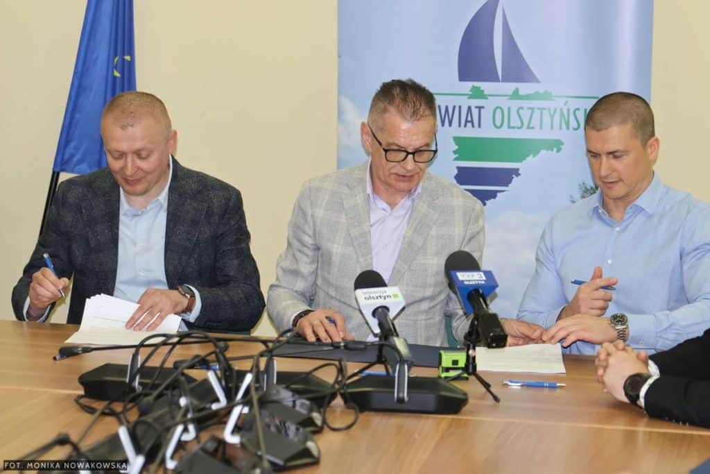 Trzech mężczyzn podpisuje umowy na inwestycje drogowe podczas konferencji prasowej.