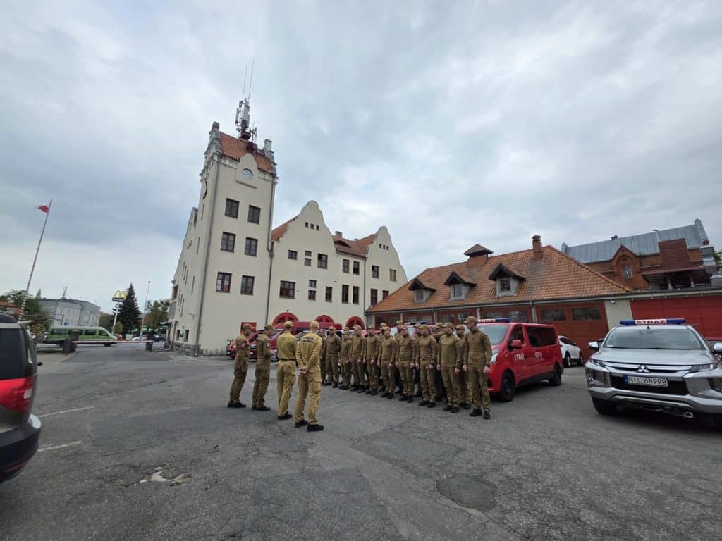 Grupa strażak&oacute;w w mundurach stoi obok pojazd&oacute;w przed budynkiem, czekając na dalsze działania.